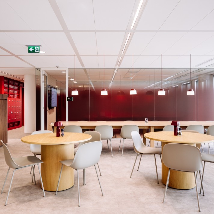 A modern office lounge area with round wooden tables and light-colored chairs. A glass wall with a red backdrop and hanging pendant lights is visible in the background, while red lockers line the left side. The space has a clean ceiling with recessed ligh