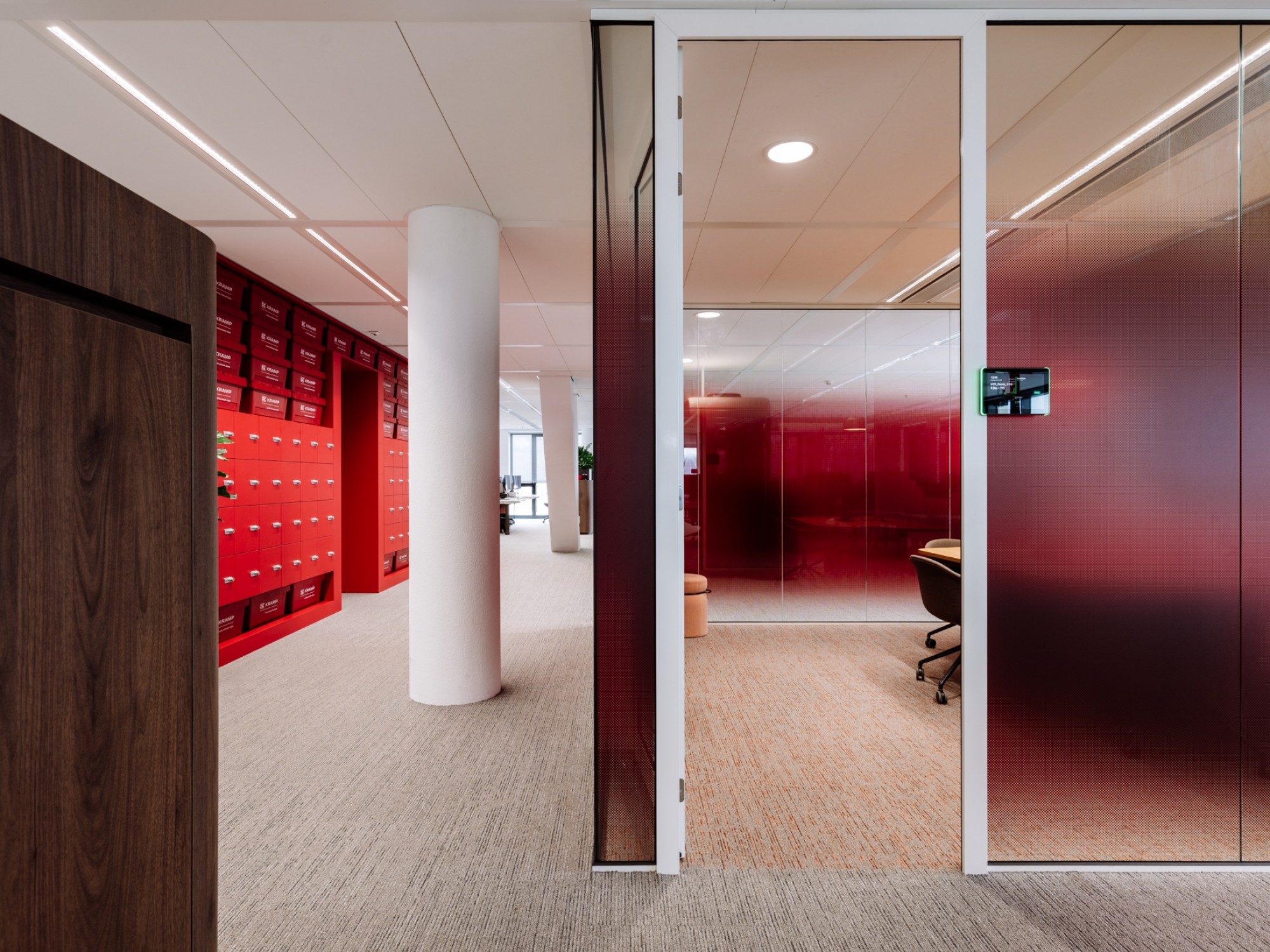 Een moderne kantoorruimte met een gang waarin rode lockers langs de muur staan. Rechts bevindt zich een glazen wand met een rood getinte afwerking en daarachter een vergaderruimte met bureaustoelen. De ruimte heeft een lichte vloer en een strak afgewerkt