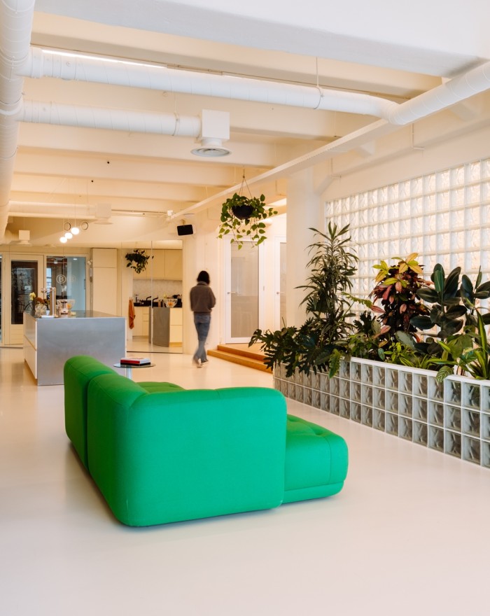 Moderne kantoorruimte met groene bank, plantenwand en open indeling, terwijl een persoon door de ruimte loopt.