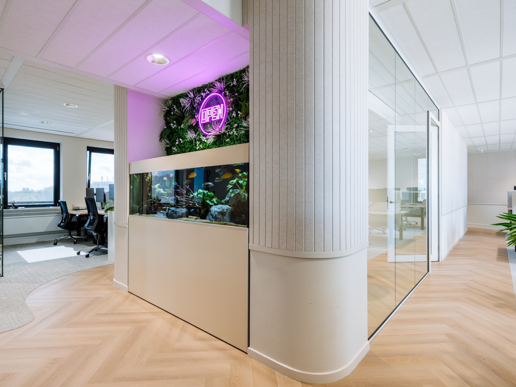 A modern office interior featuring a built-in aquarium set into a light-colored wall. Above the aquarium is a green plant wall with purple neon lighting. To the right is a glass meeting room, and the floor has a light herringbone wood pattern.