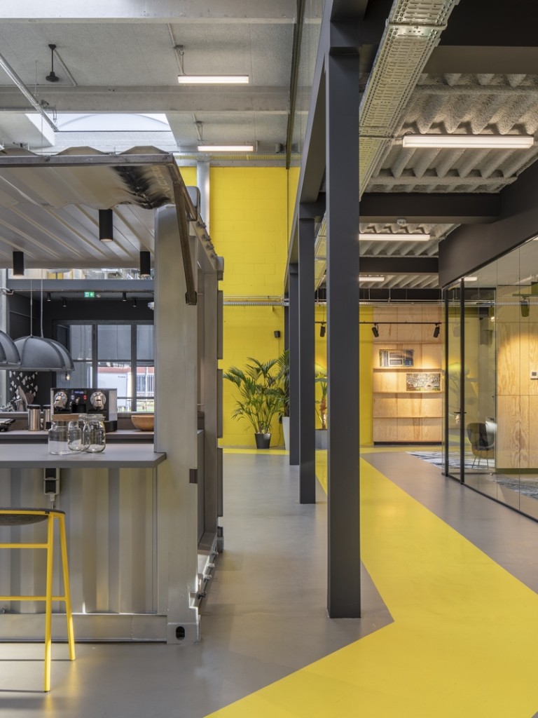 Modern kantoorinterieur met een koffiebar en gele barkrukken, glazen vergaderruimtes en een opvallend geel pad op de vloer.