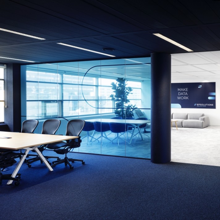 Een moderne kantoorruimte met een vergadertafel en stoelen op een donker tapijt. Rechts bevindt zich een glazen vergaderruimte met blauwe tint. Op de achterwand staat de tekst “MAKE DATA WORK” boven een lichte zithoek. De ruimte heeft een strak plafond me
