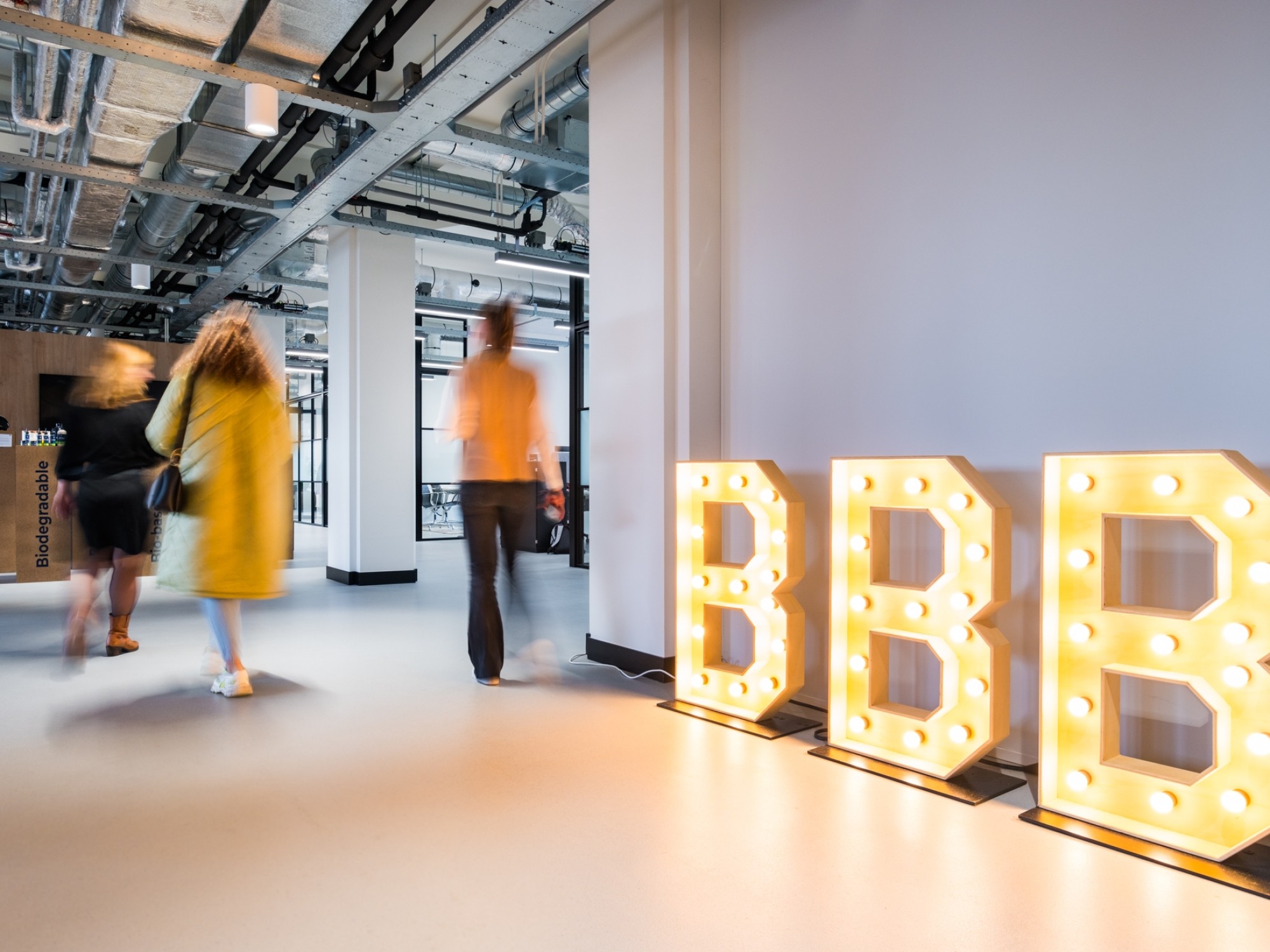 A modern office hallway with an industrial ceiling and exposed pipes. Three people walk through the space while large illuminated letters spelling “BBB” stand against the wall, glowing warmly.
