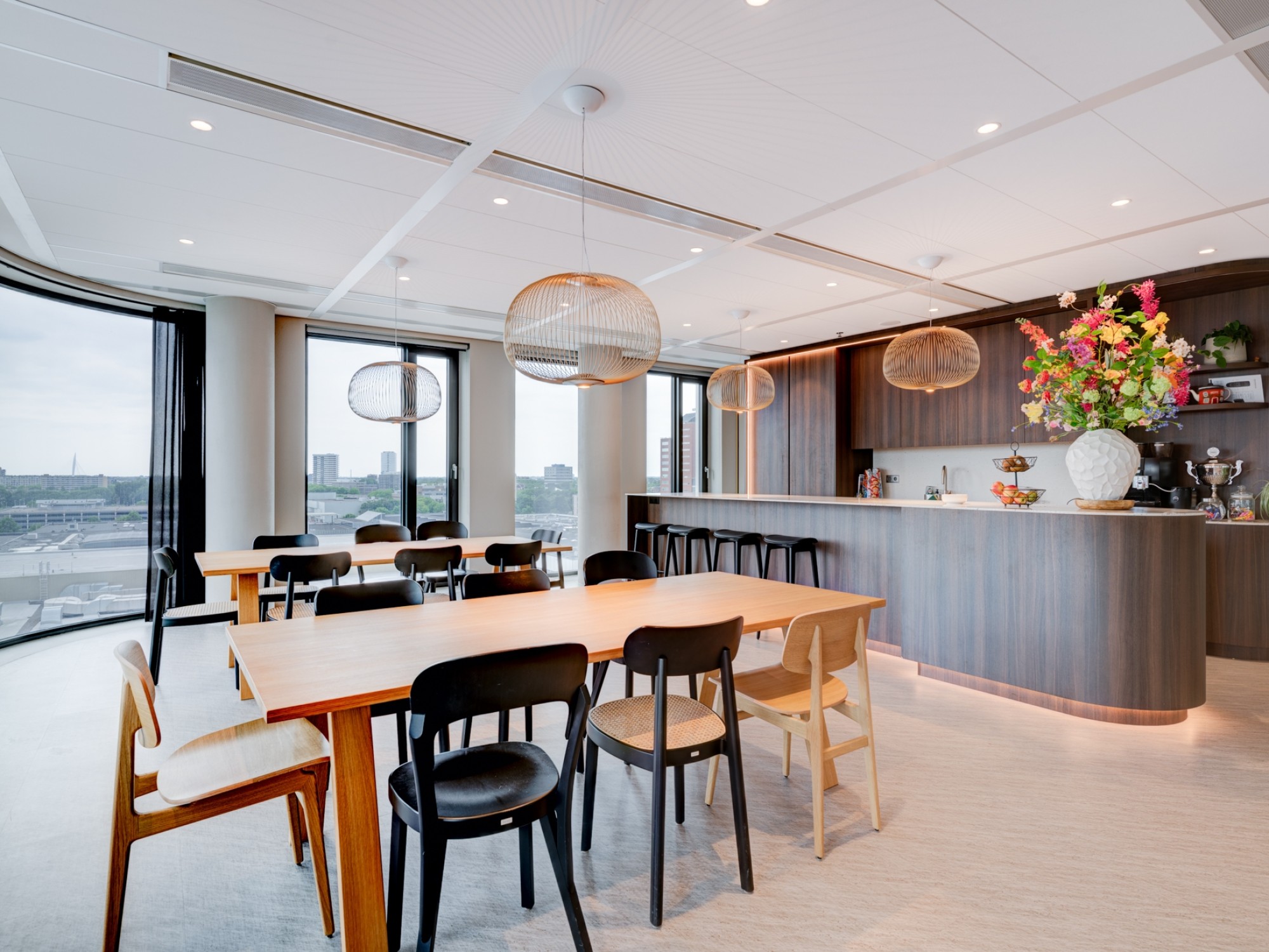 Moderne kantoorkeuken met een lange houten tafel en stoelen, bar met bloemenarrangement, hanglampen en grote ramen met uitzicht op de stad.