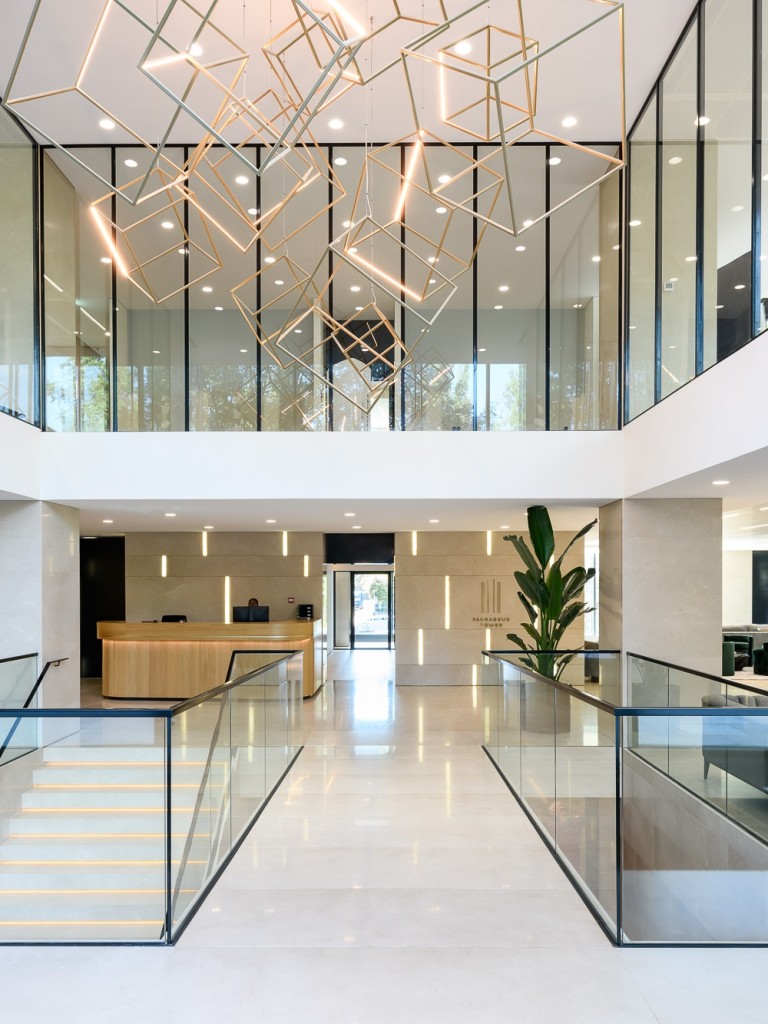 Moderne kantoorentree met glazen balustrades, open vide, design verlichting aan het plafond en planten in een lichte, ruime hal.