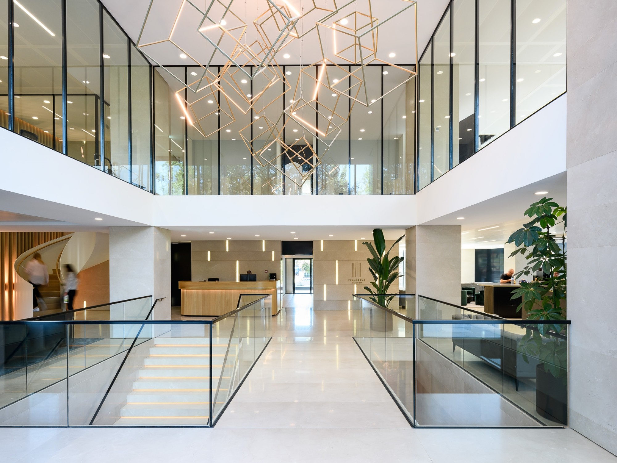 Moderne kantoorentree met glazen balustrades, open vide, design verlichting aan het plafond en planten in een lichte, ruime hal.
