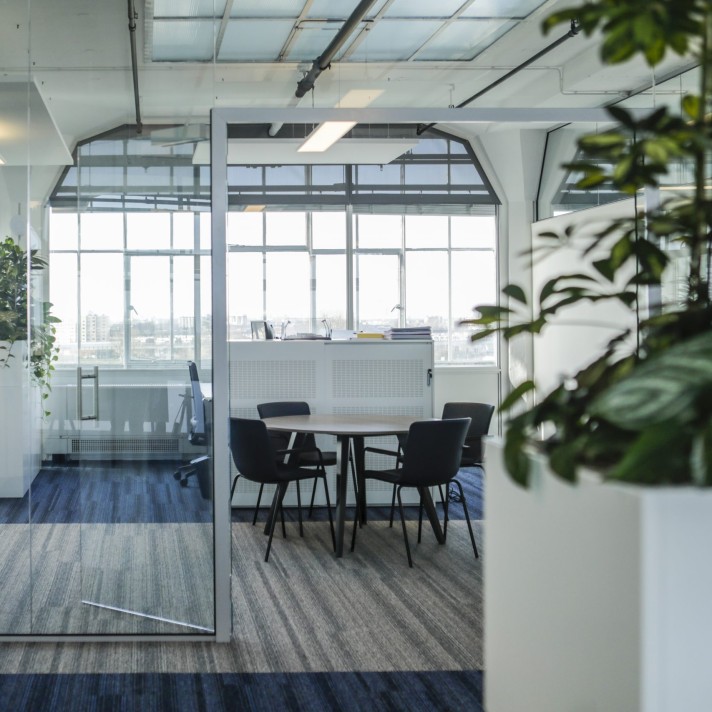 Een moderne kantoorruimte met een glazen vergaderruimte waarin een ronde tafel en vier stoelen staan. Grote ramen zorgen voor veel daglicht en uitzicht naar buiten. Op de voorgrond is een plant in een witte pot zichtbaar en het plafond heeft een industrië
