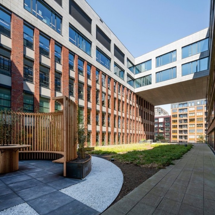 Een moderne binnentuin omringd door een kantoorgebouw met een gevel van baksteen en grote ramen. In de tuin staan planten en een houten bank met een gebogen rugleuning. De gebouwen vormen een open binnenplaats met een doorgang naar een ander complex.
