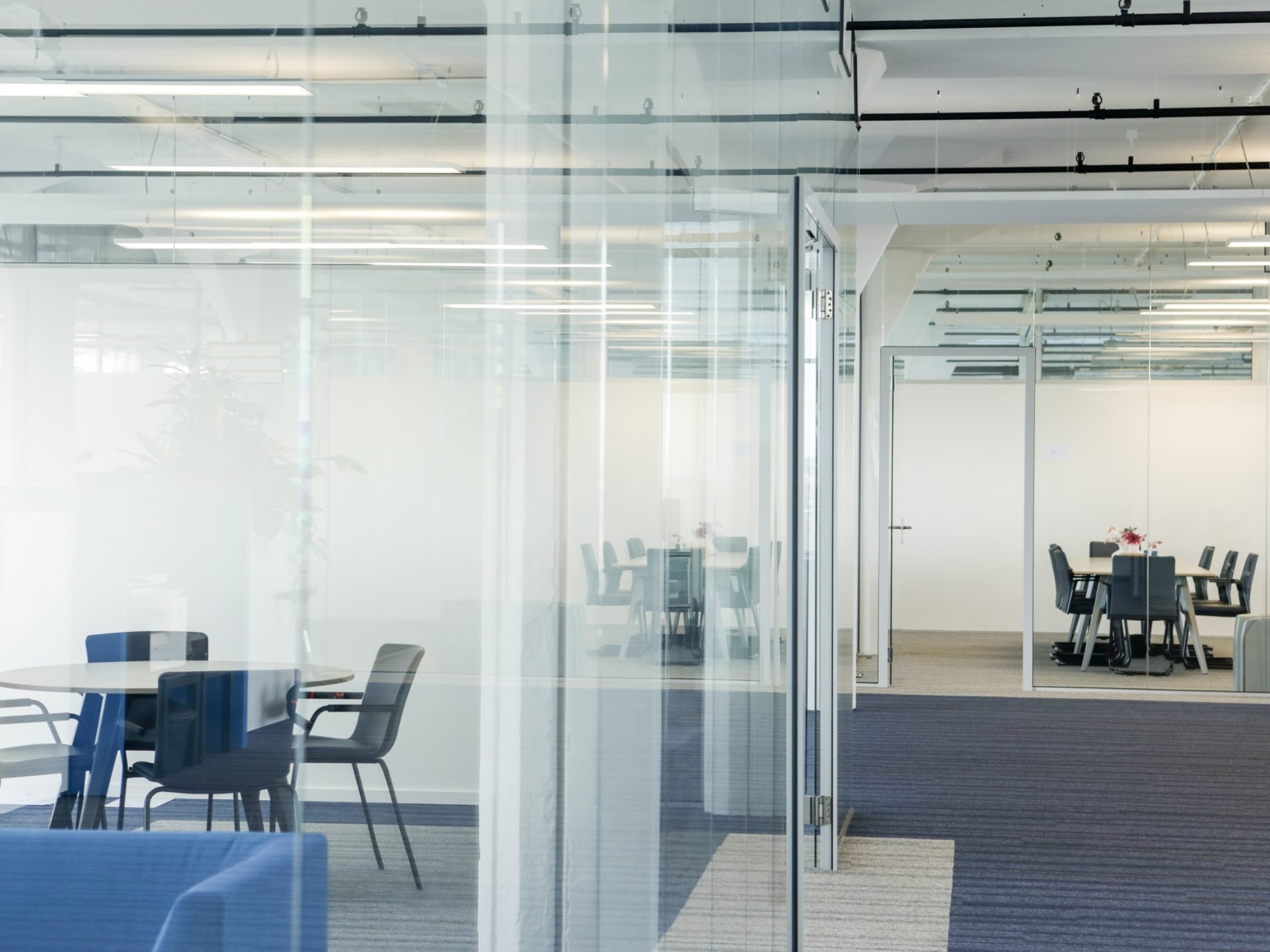 Een moderne kantoorruimte met glazen wanden en meerdere vergaderruimtes. Binnen staan tafels met stoelen op een tapijtvloer. Het plafond heeft een industriële afwerking met zichtbare leidingen en verlichting.