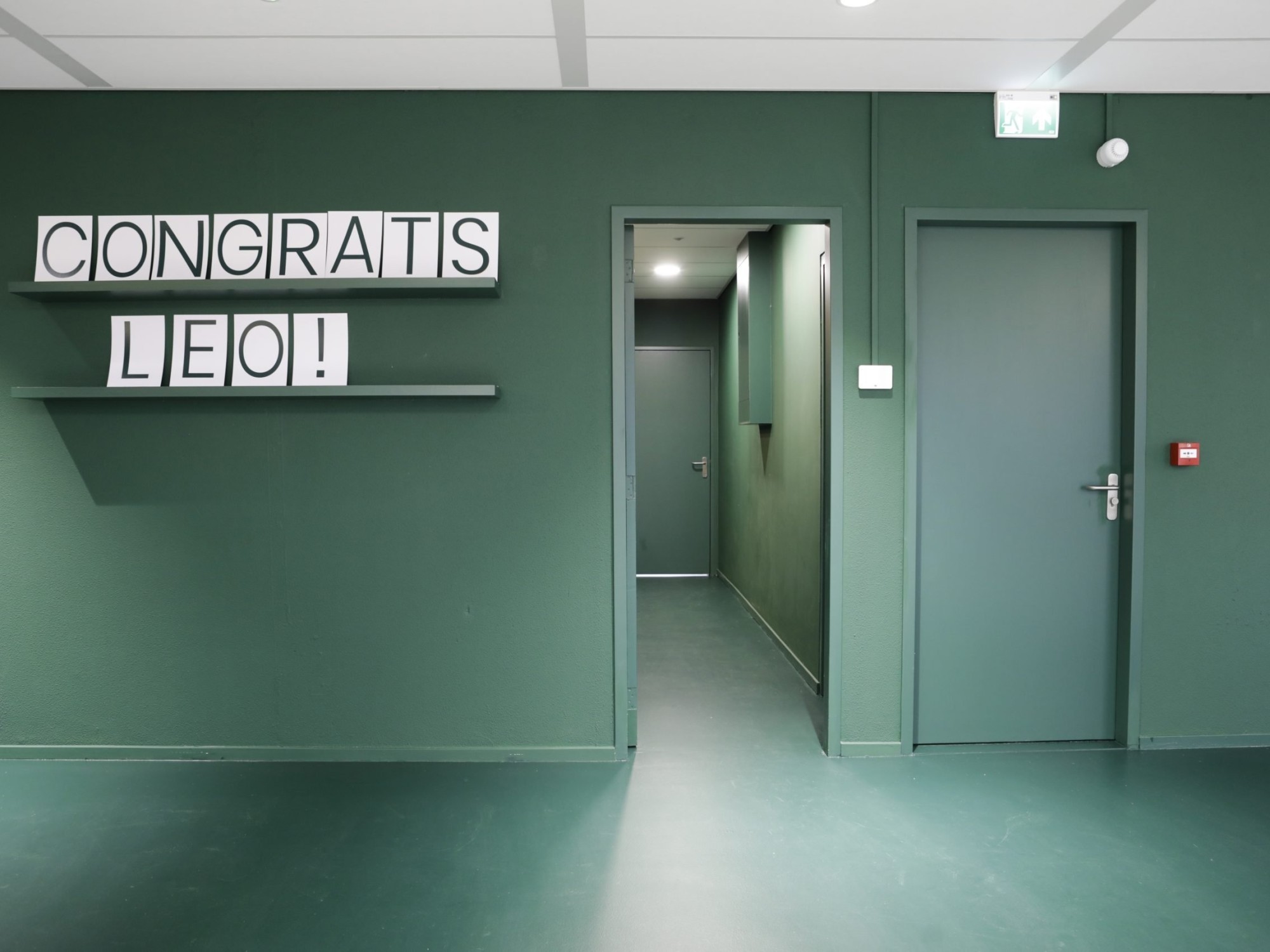 Een groene gang in een kantoorruimte met twee deuren aan de rechterzijde. Aan de linkerwand hangt een plank met blokletters waarop “CONGRATS LEO!” staat. De vloer en muren zijn in dezelfde groene tint afgewerkt.