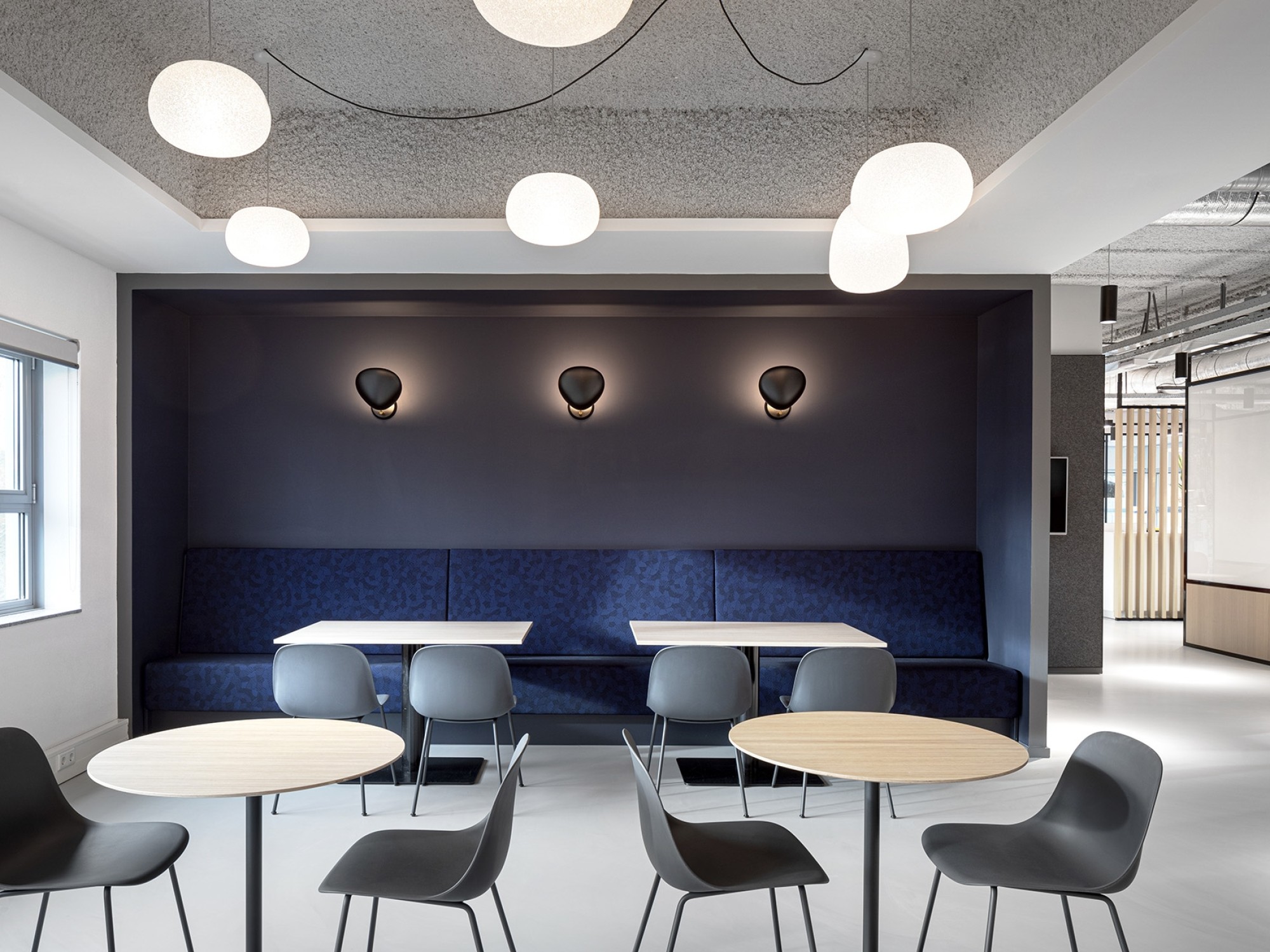 Contemporary office lounge area with round tables, gray chairs, a built-in blue upholstered bench, modern wall sconces, and large circular ceiling lights.