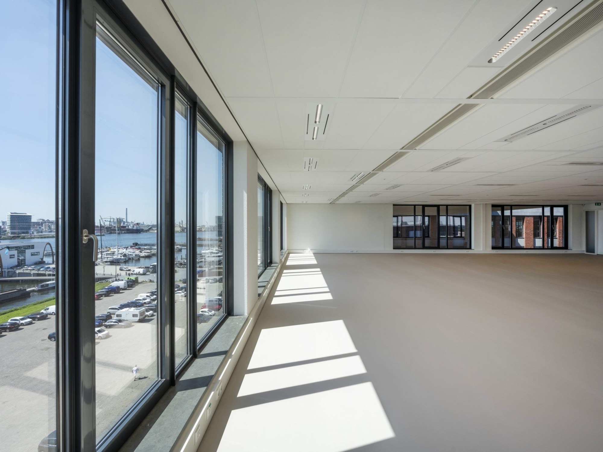 Een grote, lege kantoorruimte met vloer-tot-plafondramen langs één zijde. Zonlicht valt naar binnen en werpt lange schaduwen op de vloer. Door de ramen is uitzicht op een parkeerplaats en omliggende gebouwen zichtbaar. De ruimte heeft een strak plafond me