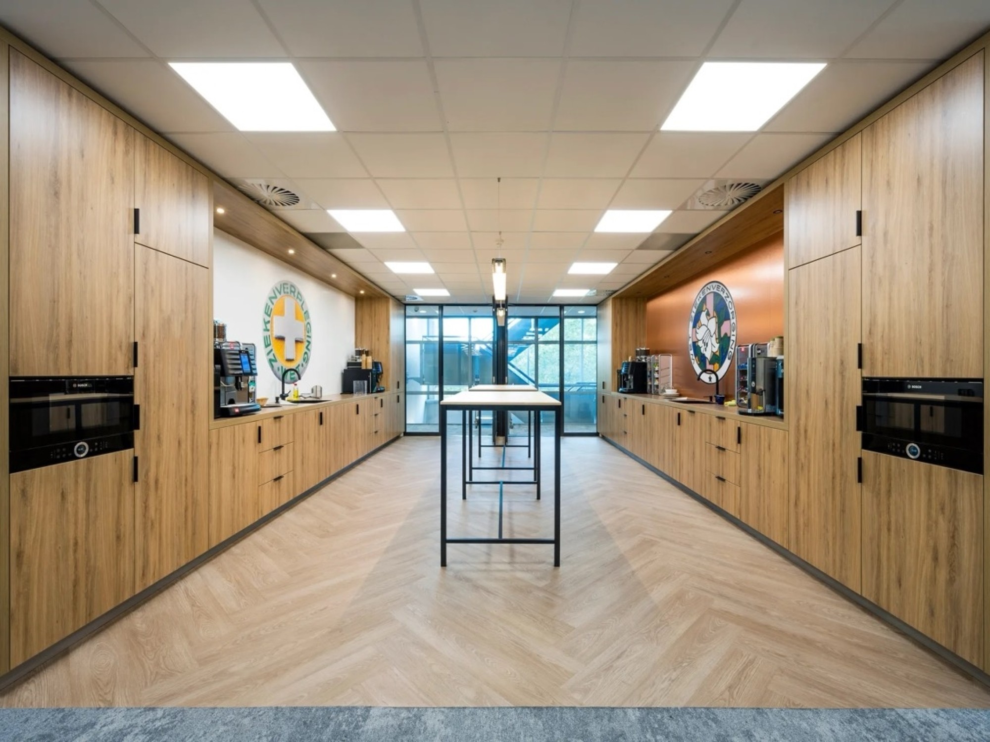 Modern office kitchen with wooden cabinets on both sides, built-in coffee machines, a central high table, and large windows at the end of the room.