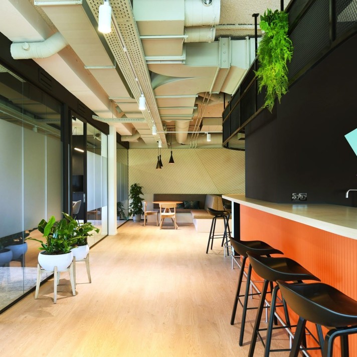 Een moderne kantoorruimte met een lange bar met zwarte barkrukken en een oranje accent aan de onderzijde. Links bevinden zich glazen vergaderruimtes en planten in potten. Het plafond heeft een industriële afwerking met zichtbare leidingen en verlichting.