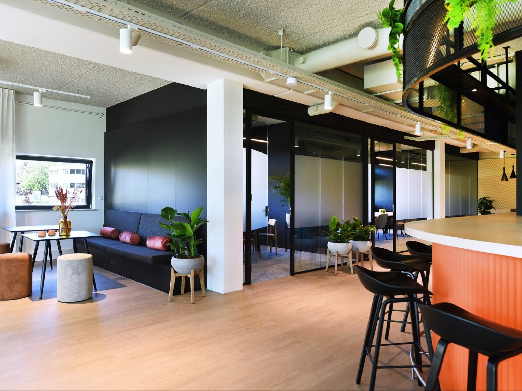 Een moderne kantoorruimte met een bar met zwarte barkrukken en een oranje accent. Links bevindt zich een zithoek met een donkere bank en poefjes bij een raam. In het midden zijn glazen vergaderruimtes met zwarte frames zichtbaar. Het plafond heeft een ind