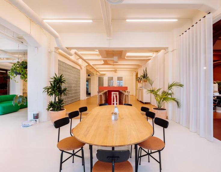 Moderne, lichte vergaderruimte met een lange houten tafel, zwarte stoelen, groene planten en witte gordijnen in een open kantooromgeving.