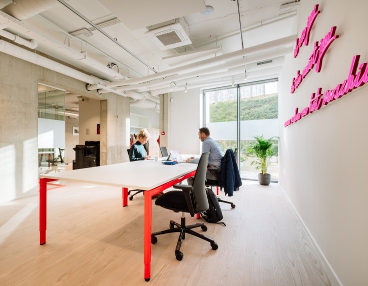 Lichte, moderne kantoorruimte met grote tafel op rode poten en medewerkers in overleg bij het raam, met roze wandtekst op de muur.