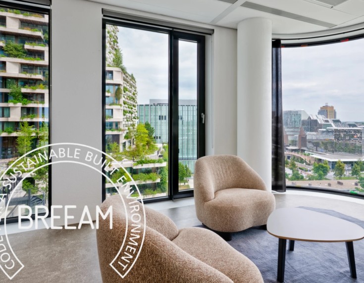 Modern lounge area with comfortable seating, large windows, and a city view featuring green residential buildings and urban surroundings.