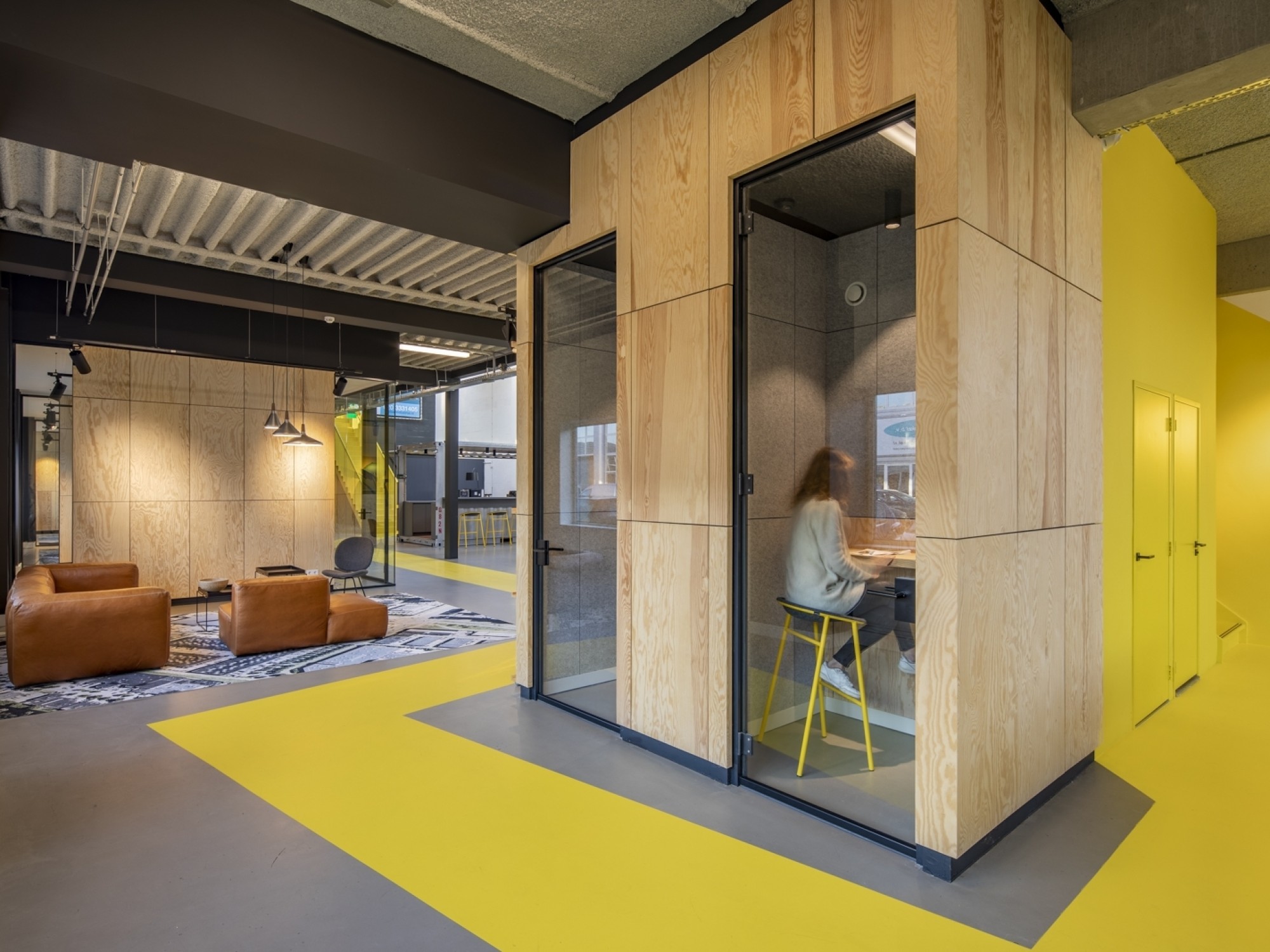 Urban industrial kantoor met gele looproutes, houten belcellen en loungehoek met leren fauteuils