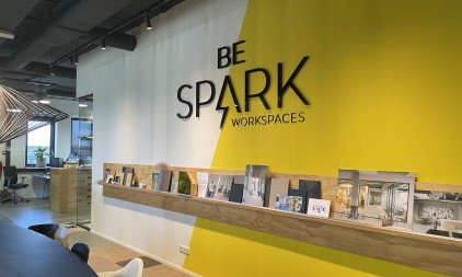 Interior of a modern coworking space with “Be Spark Workspaces” sign on a yellow wall and a display shelf with magazines.