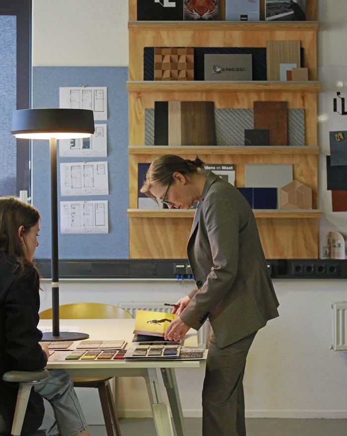 Twee personen bekijken materiaalstalen aan een bureau, met een wand vol voorbeelden en kleurenstalen op de achtergrond in een ontwerpstudio.