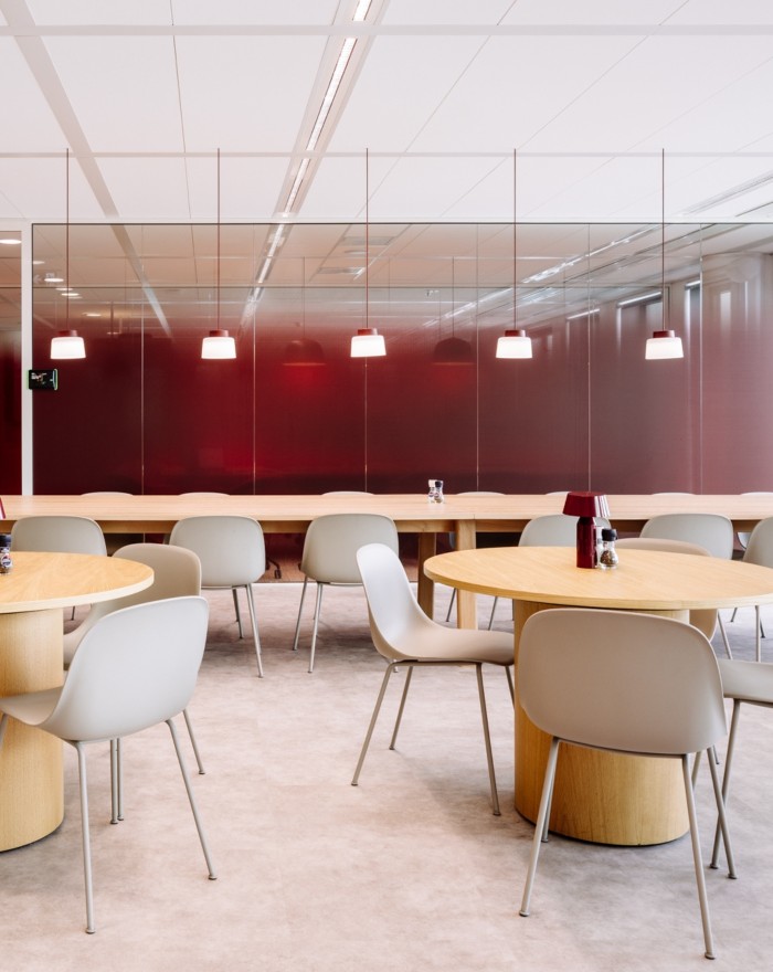 Moderne kantine met ronde tafels, lichte stoelen en glazen wand met rode achtergrond, sfeervol verlicht door hanglampen.