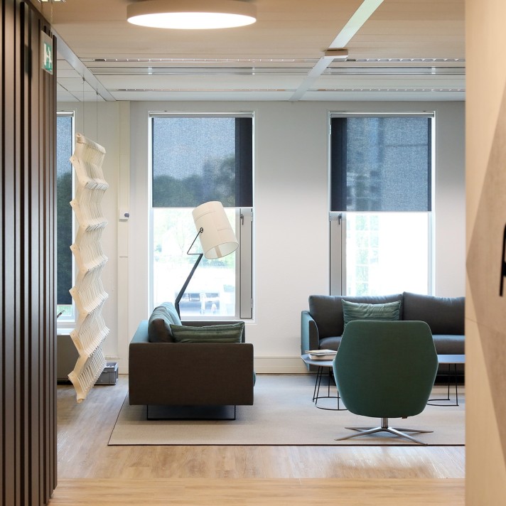 Een moderne zithoek in een kantoor met twee donkere banken en een groene fauteuil rondom een klein rond tafeltje. Grote ramen met half gesloten rolgordijnen zorgen voor daglicht. Links staat een opvallende witte designlamp en de ruimte heeft een lichte ho