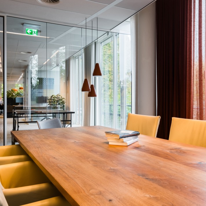 Een moderne vergaderruimte met een grote houten tafel en gele stoelen. Op de tafel liggen enkele boeken. Naast de tafel hangen drie zwarte hanglampen. Achter de glazen wand is een andere vergaderruimte zichtbaar en grote ramen zorgen voor veel natuurlijk