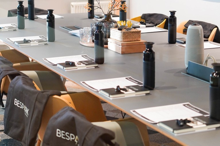 Vergadertafel met notitieblokken, waterflessen en stoelen met ‘BESPARK’ op de rug in een moderne trainings- of workshopruimte.
