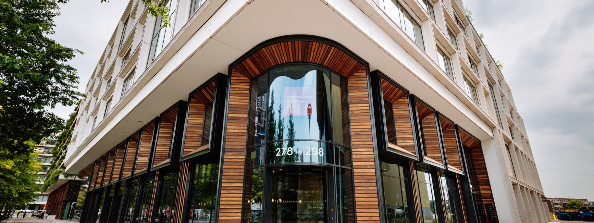 Modern kantoorgebouw met afgeronde glazen hoek, houten geveldetails en grote ramen op straatniveau.