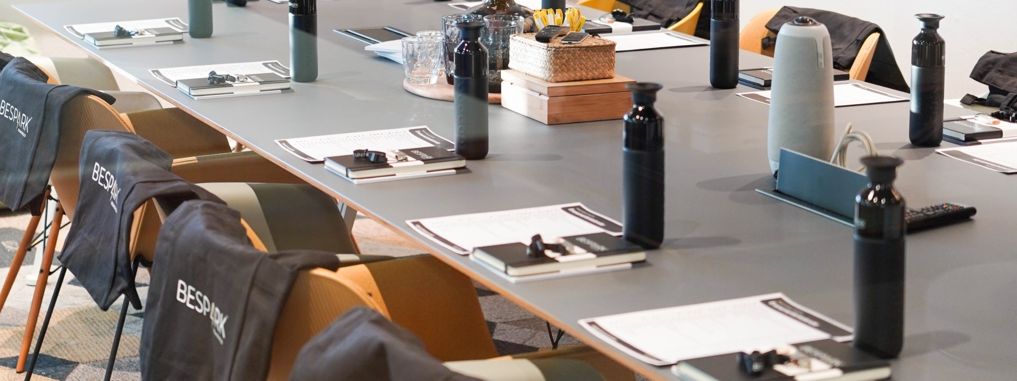 Vergadertafel met notitieblokken, waterflessen en stoelen met ‘BESPARK’ op de rug in een moderne trainings- of workshopruimte.