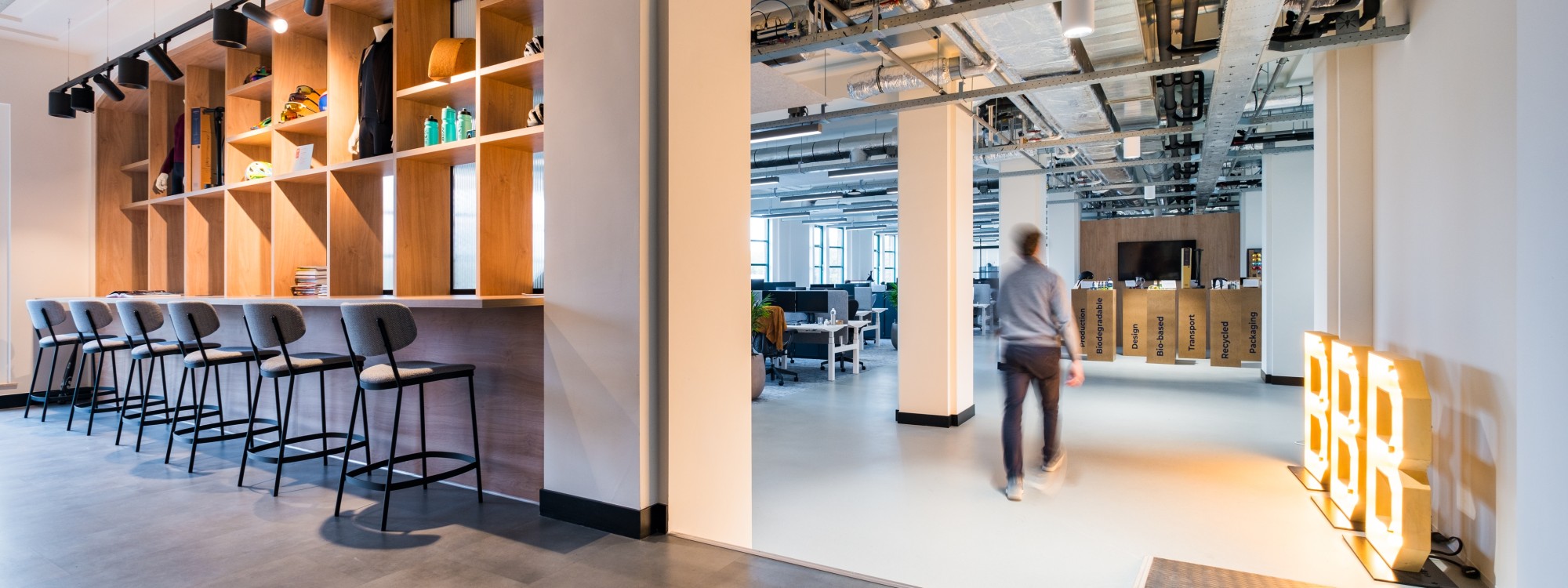 Modern office interior with bar seating, open workspace, industrial ceiling details, and a person walking through the space.