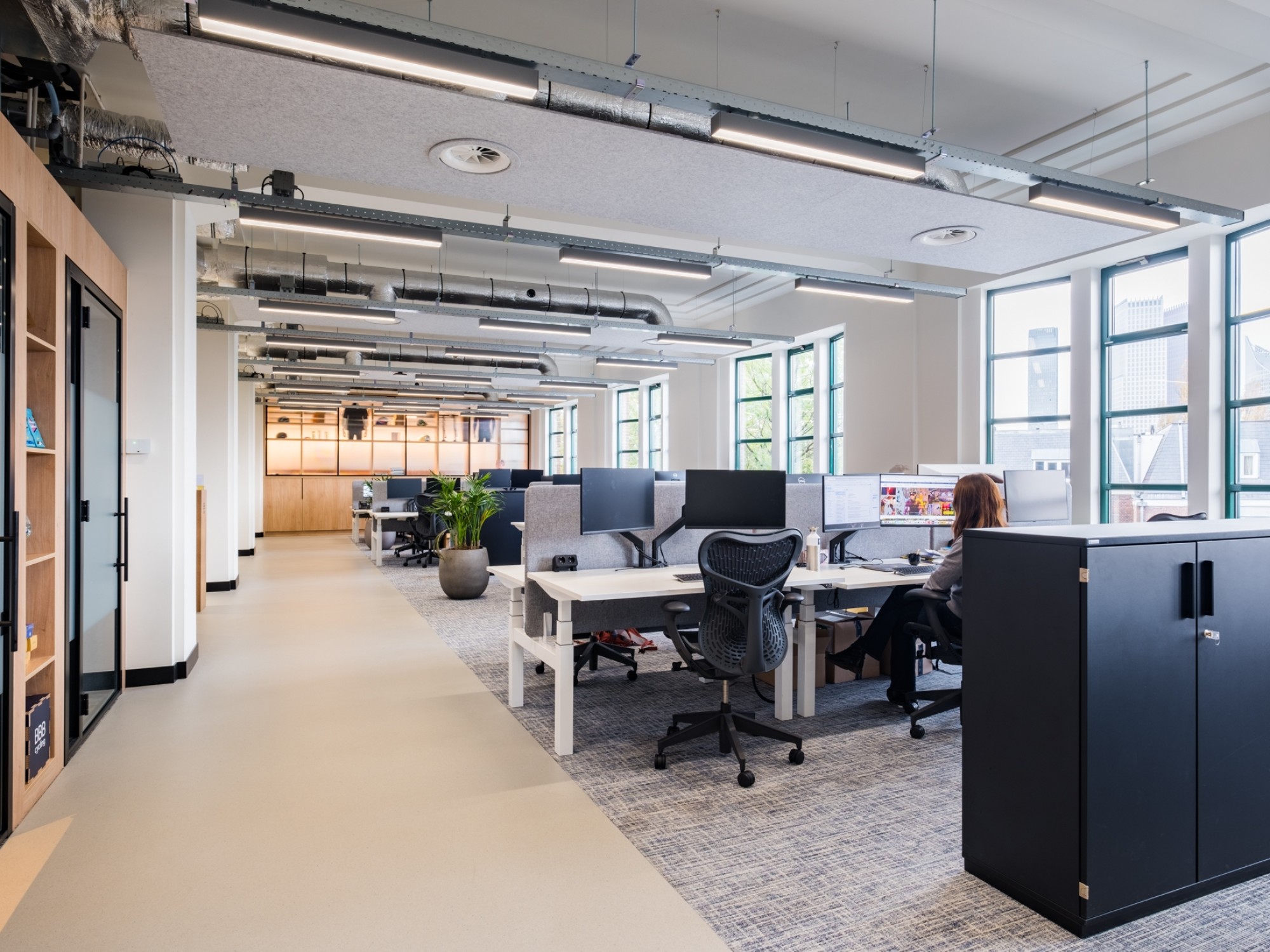 Open kantoorruimte met bureaus, ergonomische stoelen en grote ramen die zorgen voor veel daglicht, met medewerkers aan het werk.