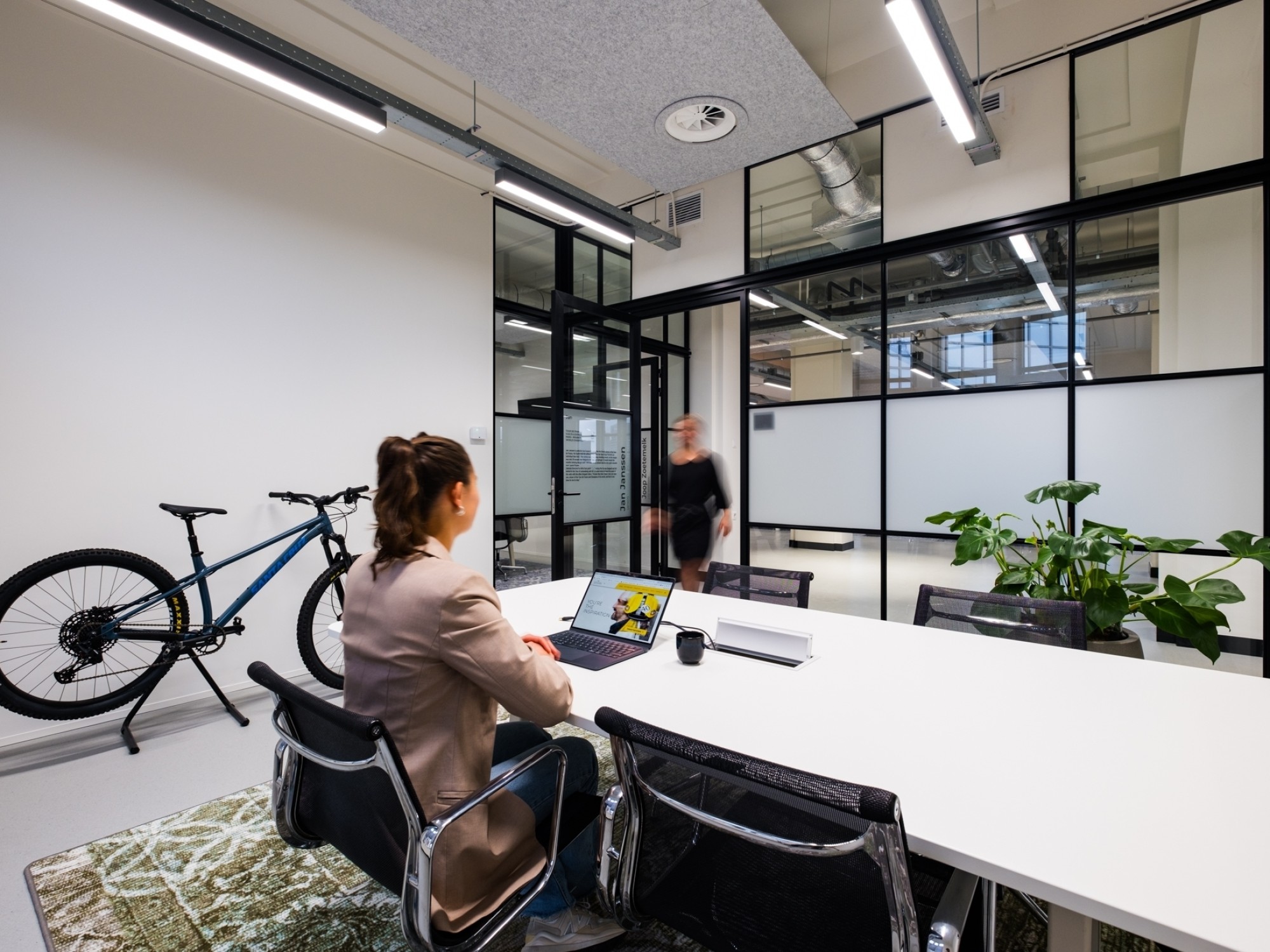 Moderne vergaderruimte met glazen wanden en industriële verlichting. Een persoon zit aan een witte vergadertafel met een laptop, terwijl op de achtergrond iemand langsloopt. Aan de linkerkant staat een fiets op een standaard en rechts staan groene planten