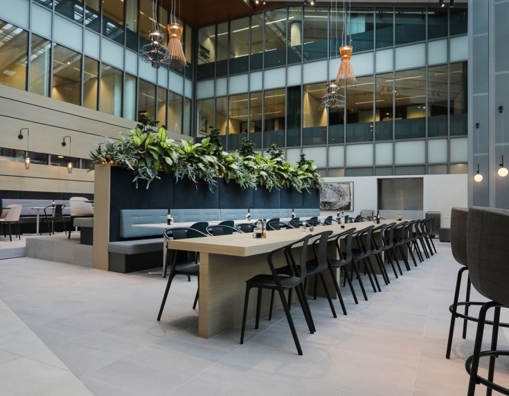 Grote open kantoorruimte met lange tafel, zitbanken en planten in een modern atrium.