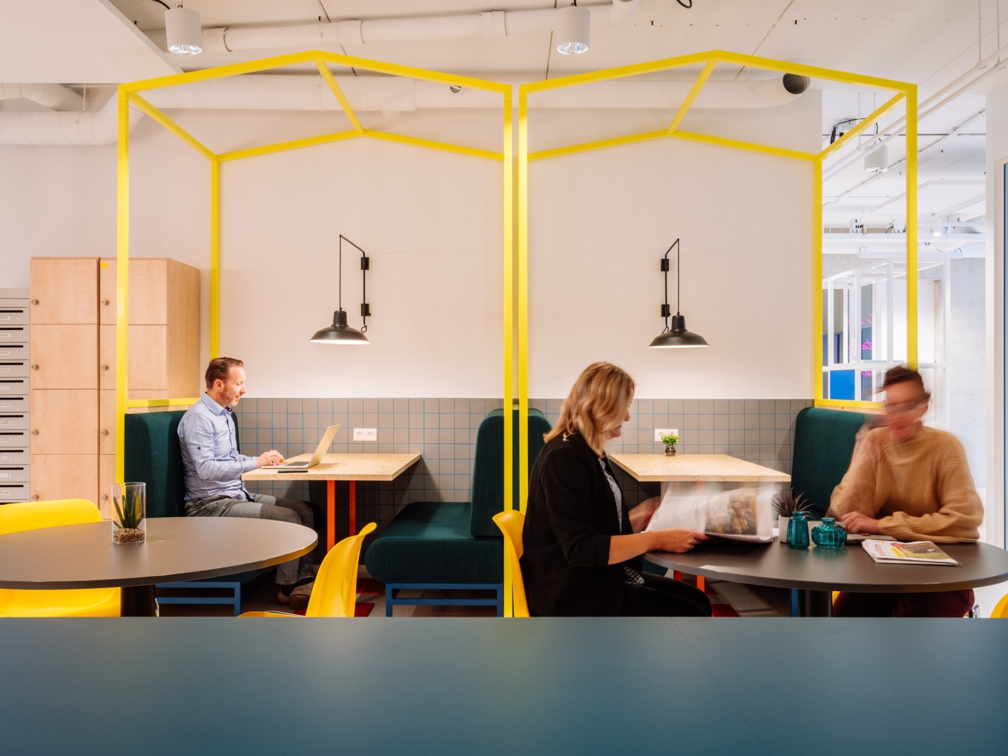 Moderne kantoor- en ontmoetingsruimte met kleurrijke zitplekken en gele frameconstructies boven de tafels. Medewerkers werken en overleggen aan ronde tafels en in zitnissen, wat een informele en creatieve werksfeer uitstraalt.