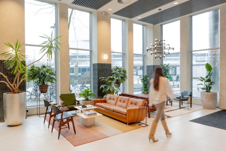 Lichte kantoorlounge met grote ramen, veel planten en een leren bank, terwijl een persoon door de ruimte loopt.