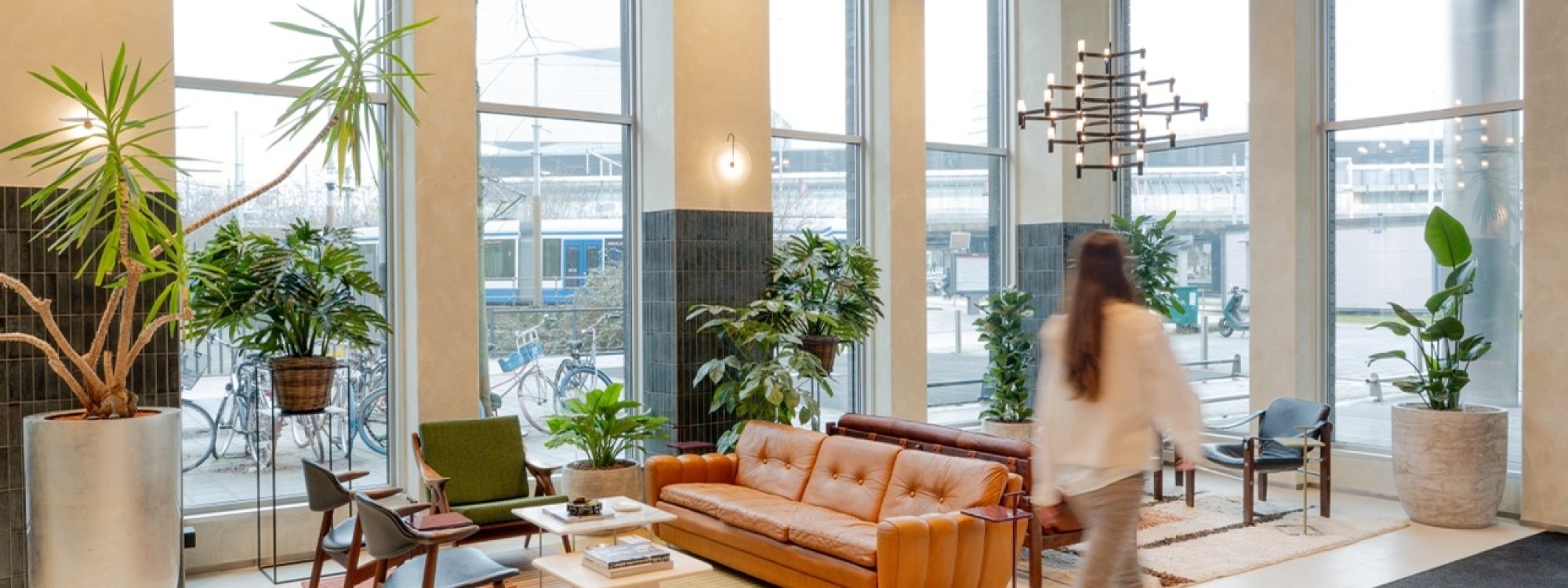 Lichte kantoorlounge met grote ramen, veel planten en een leren bank, terwijl een persoon door de ruimte loopt.