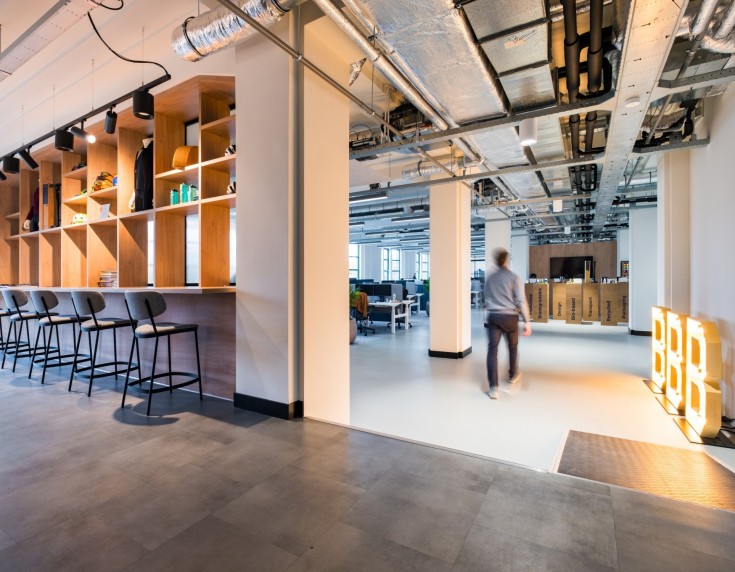 Modern kantoorinterieur met bar en hoge krukken, open werkruimte met industriële plafonds en een persoon die door de ruimte loopt, met tekstoverlay ‘Het kantoor van de toekomst’.