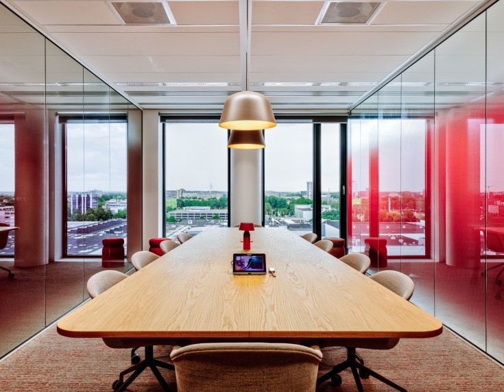 Moderne vergaderruimte met een lange houten tafel en beige stoelen, omringd door glazen wanden met rode accenten, met uitzicht op een stedelijk landschap door grote ramen.