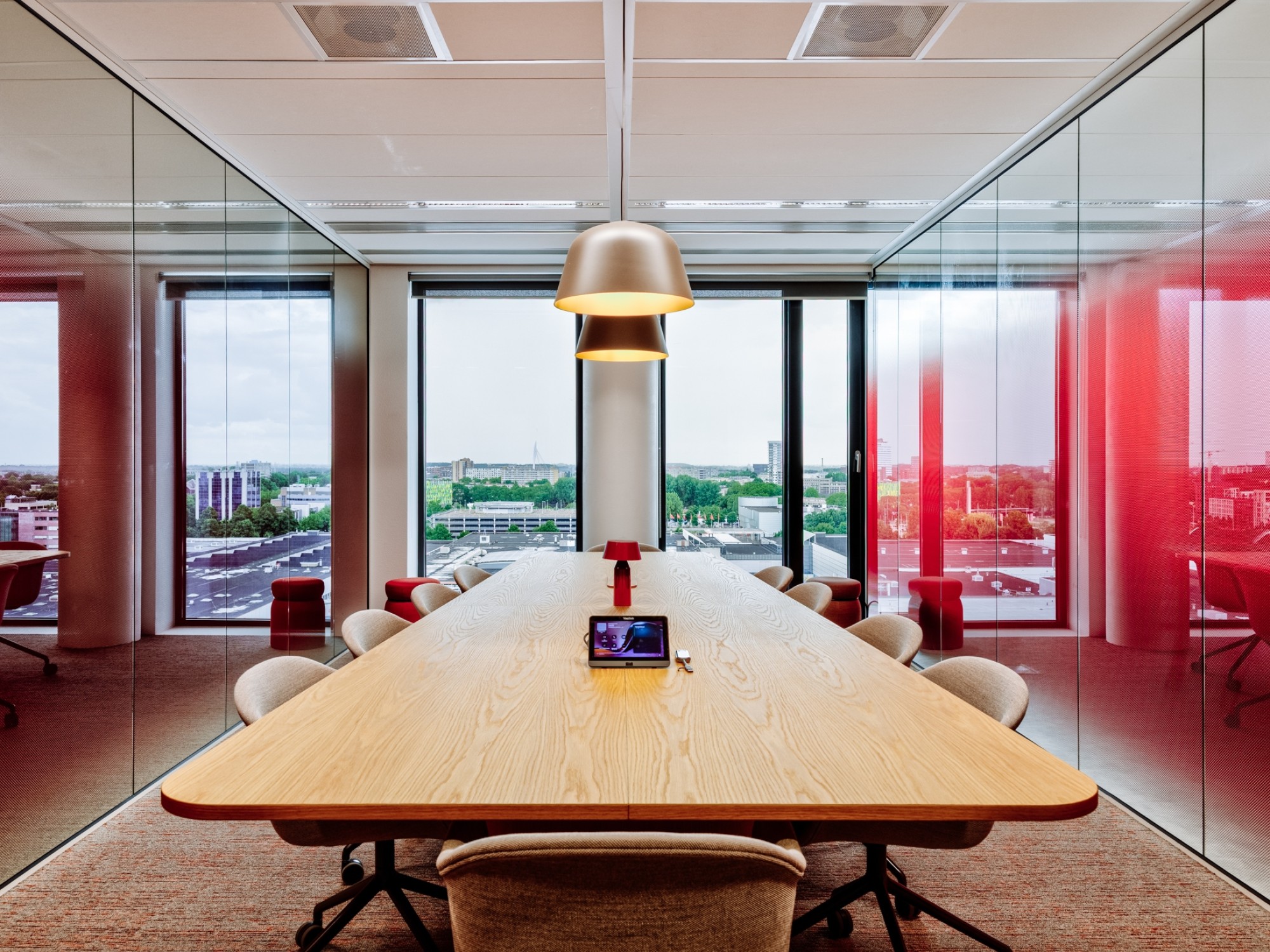 Moderne vergaderruimte met een lange houten tafel en beige stoelen, omringd door glazen wanden met rode accenten, met uitzicht op een stedelijk landschap door grote ramen.