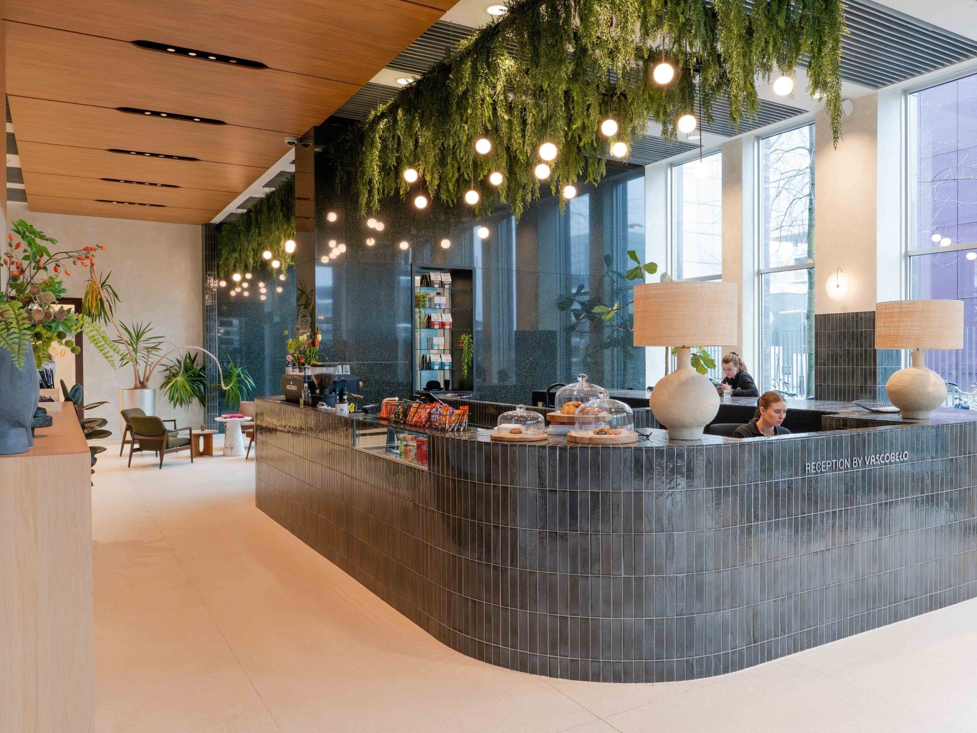 Een moderne vergaderruimte met een ronde houten tafel en bruine stoelen. Aan de muur hangt een groot kunstwerk en via de hoge ramen is uitzicht op kantoorgebouwen en groen in de stad. De ruimte is licht en strak ingericht met een rustige uitstraling.