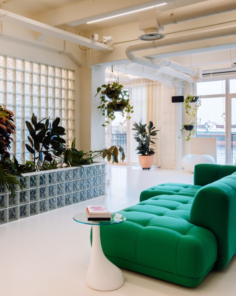Lichte kantoorruimte met groene bank, hangplanten en glazen bouwstenen wand.