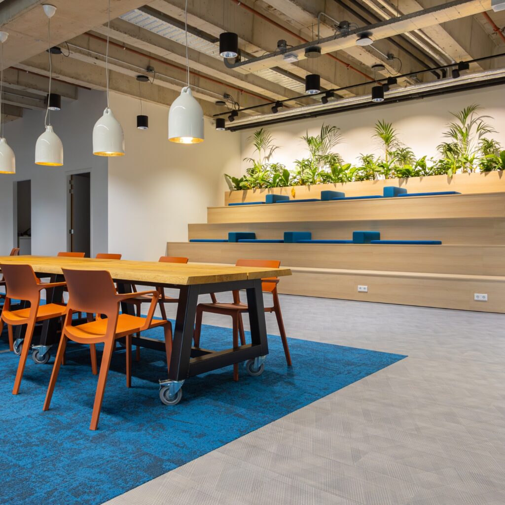 Moderne kantoorruimte met hoge tafels, oranje stoelen en houten tribune met planten voor informele bijeenkomsten.