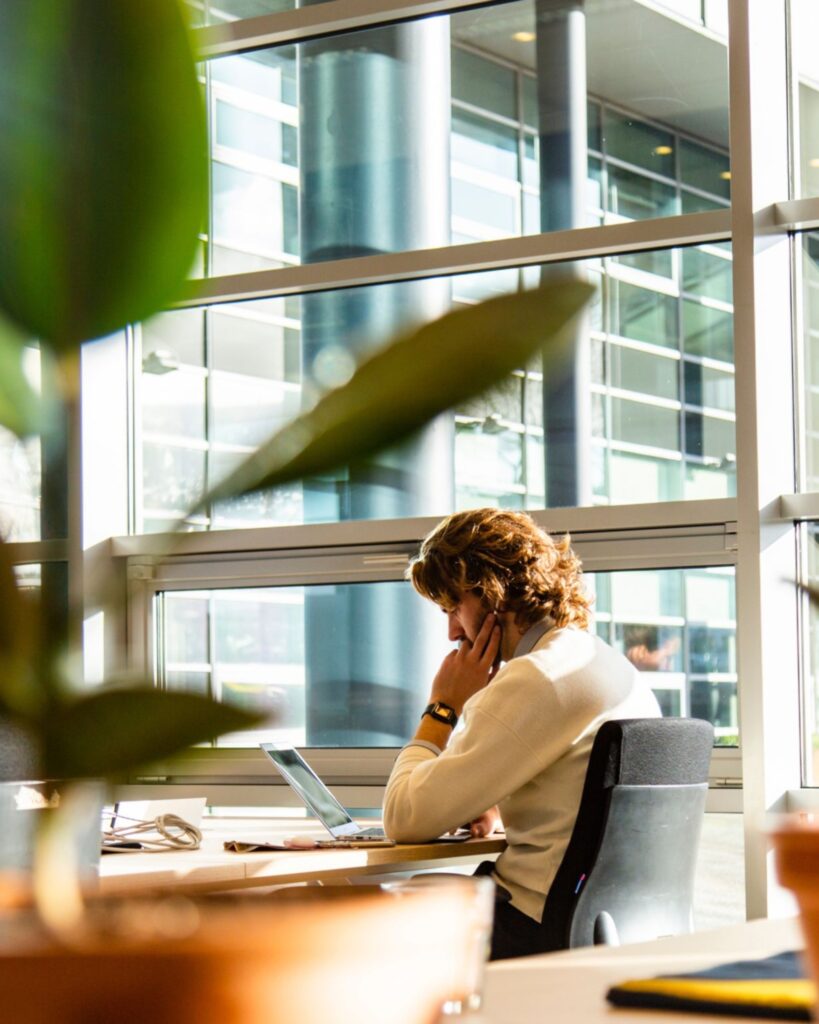Persoon werkt geconcentreerd op een laptop bij een raam met veel daglicht, omringd door een moderne kantooromgeving met groene planten op de voorgrond.