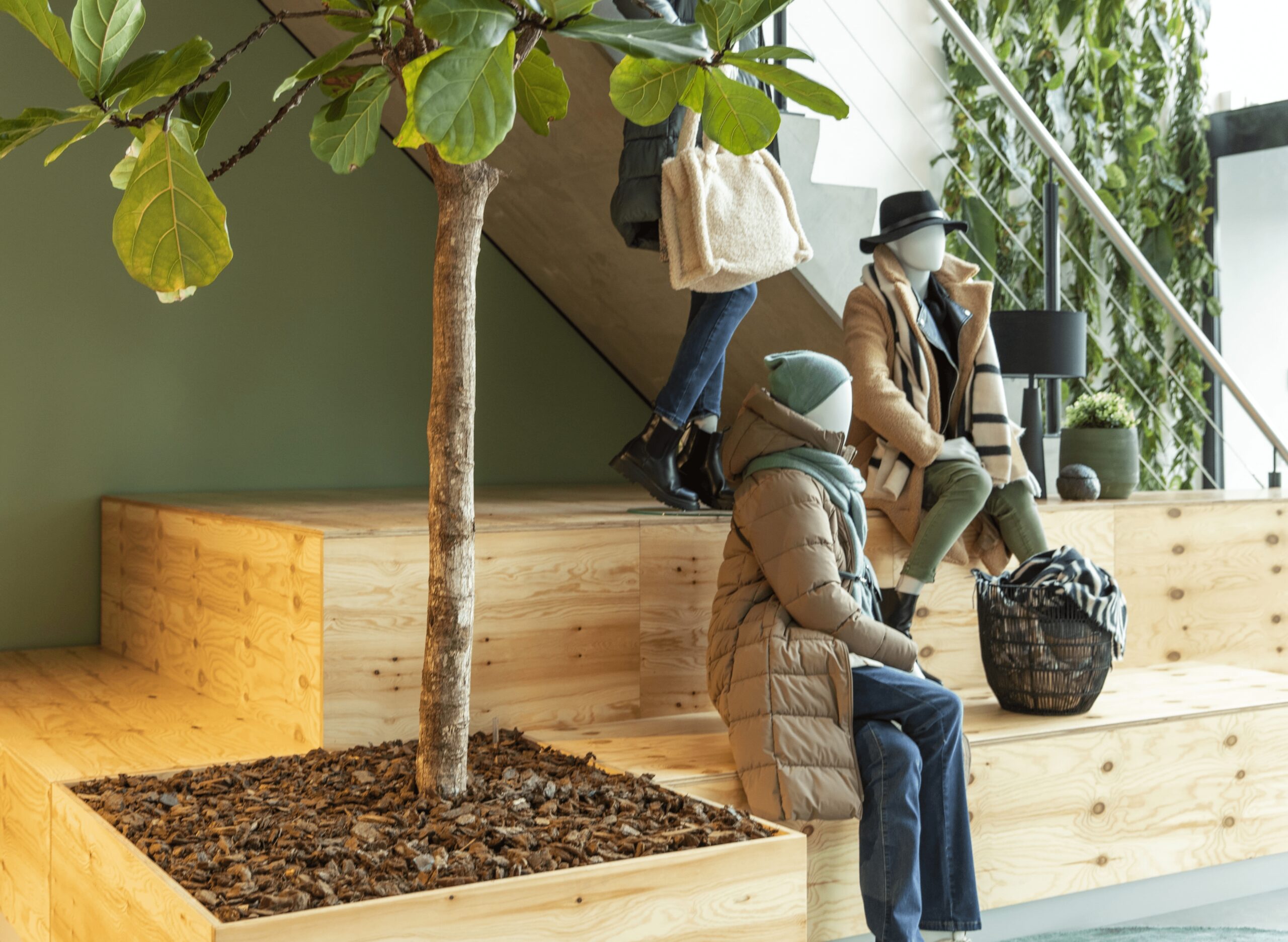 Een moderne binnenruimte met houten zitblokken rond een boom in een plantenbak. Twee personen zitten op de houten constructie, gekleed in winterjassen en mutsen, terwijl een derde persoon langsloopt. De ruimte heeft een groene wand en een lichte, open uit
