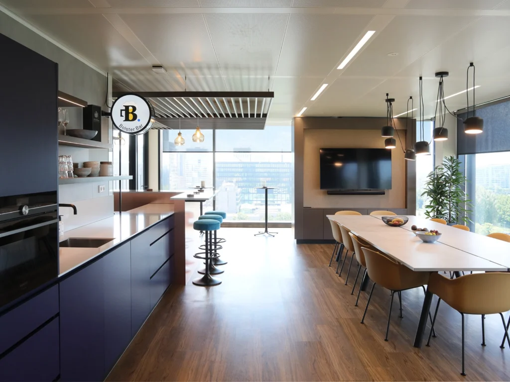 Een moderne kantoorruimte met een keuken, barstoelen en een vergadertafel met stoelen.
