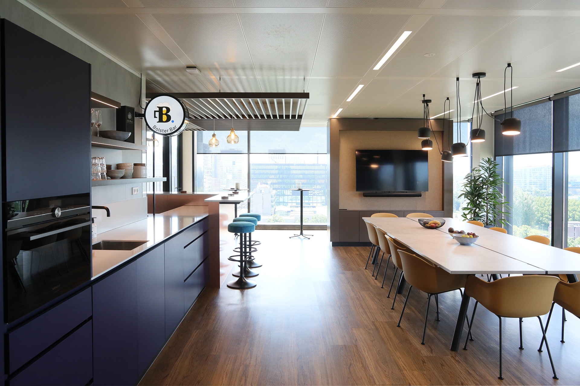Modern office break room with kitchenette, a long meeting table, and large windows.