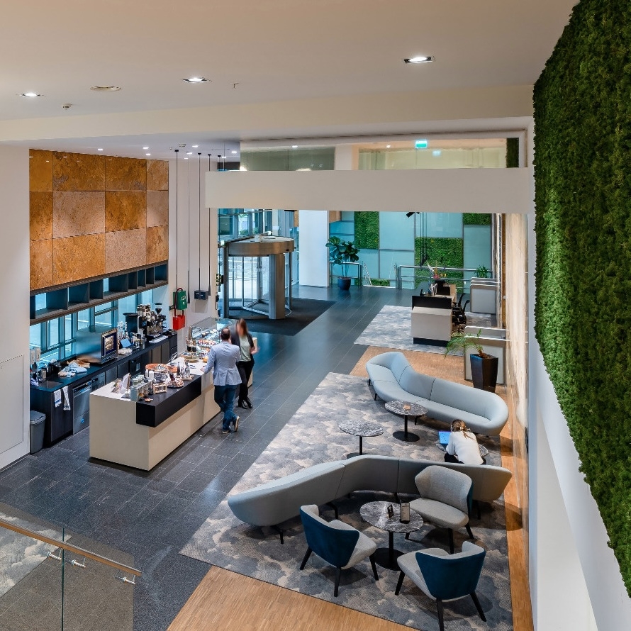 Een moderne kantoorentree met een open vide en een koffiebar aan de linkerzijde. In het midden staat een zithoek met banken en stoelen rondom lage tafeltjes. Rechts loopt een hoge groene plantenwand langs de muur. De ruimte is licht en ruim opgezet met ee