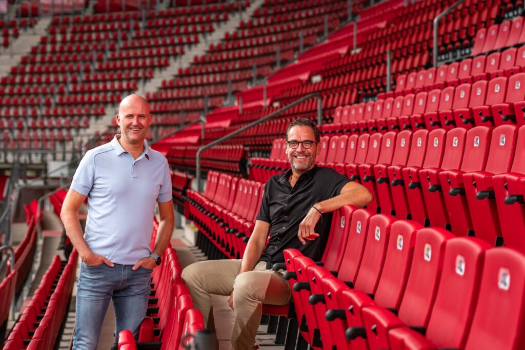 Twee mannen poseren tussen rode stadionstoelen op de tribune.